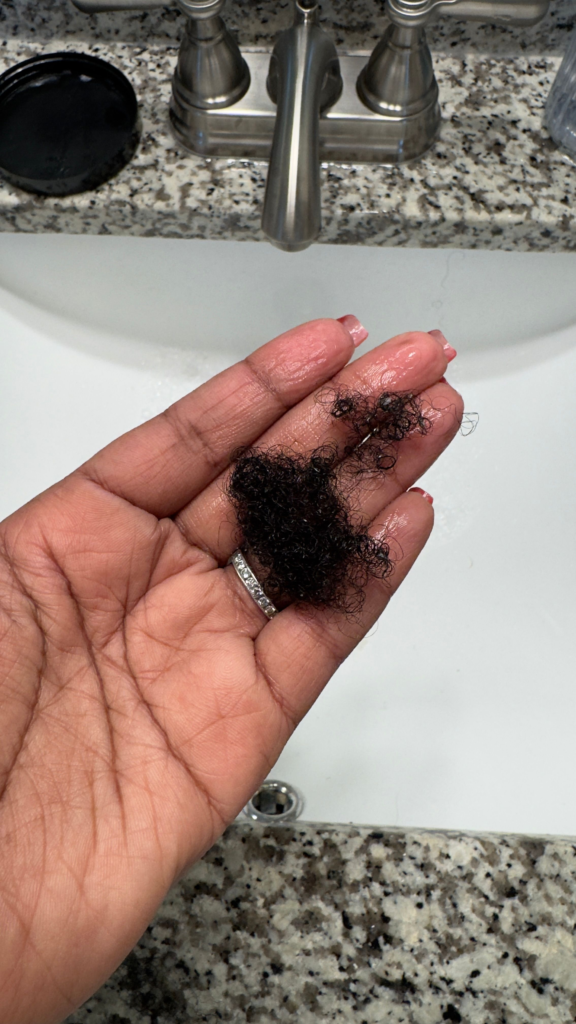 A close-up of a hand holding a small amount of shed curly hair from detangling with The Doux 808 Gel and Chief Rocka Mousse for Defined Curls wash-and-go routine. The background features a bathroom sink with a granite countertop and a stainless steel faucet.