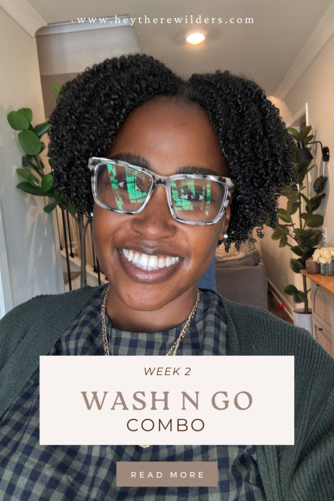 A smiling woman with naturally curly hair and glasses takes a selfie indoors. She is wearing a green cardigan over a checkered dress, with gold jewelry. The image features a text overlay that reads "Week 2 Wash N Go Combo" with a "Read More" button, and the website "www.heytherewilders.com" at the top. The background includes a cozy, well-decorated living space with plants and warm lighting.