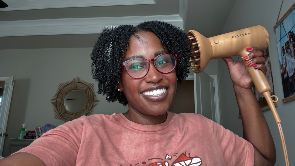 A smiling woman with defined, hydrated curls using The Doux 808 Gel and Chief Rocka Mousse for Defined Curls wash-and-go routine. She holds a gold Pattern hairdryer with a diffuser attachment, showcasing her curl definition and volume.